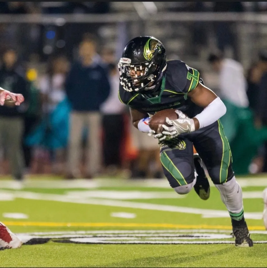 Ace in a black and green uniform carrying the football upfield during a night game.