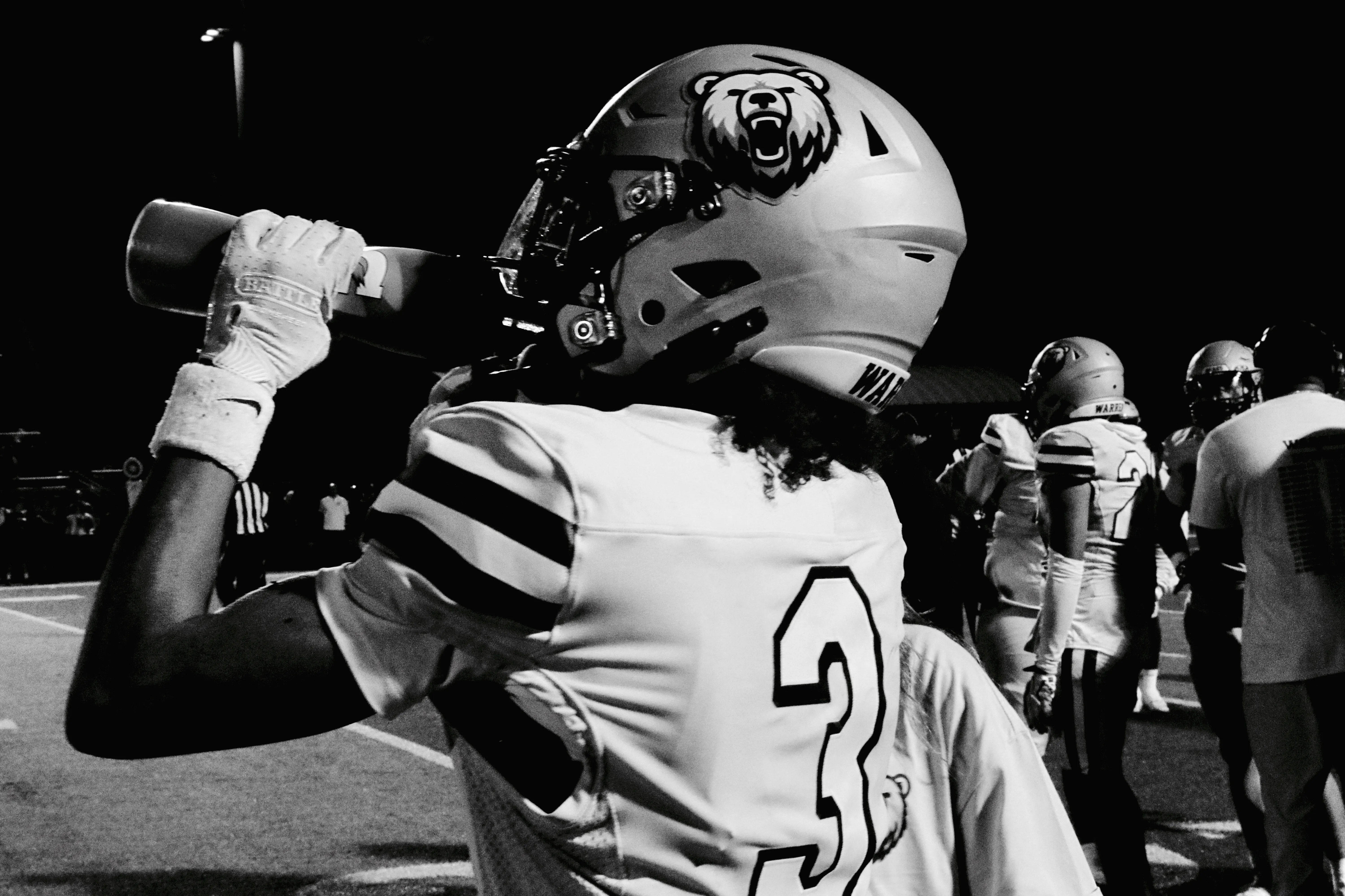 Black-and-white photo of a Warren Bears player wearing number 3, tipping back a water bottle on the sideline at night.