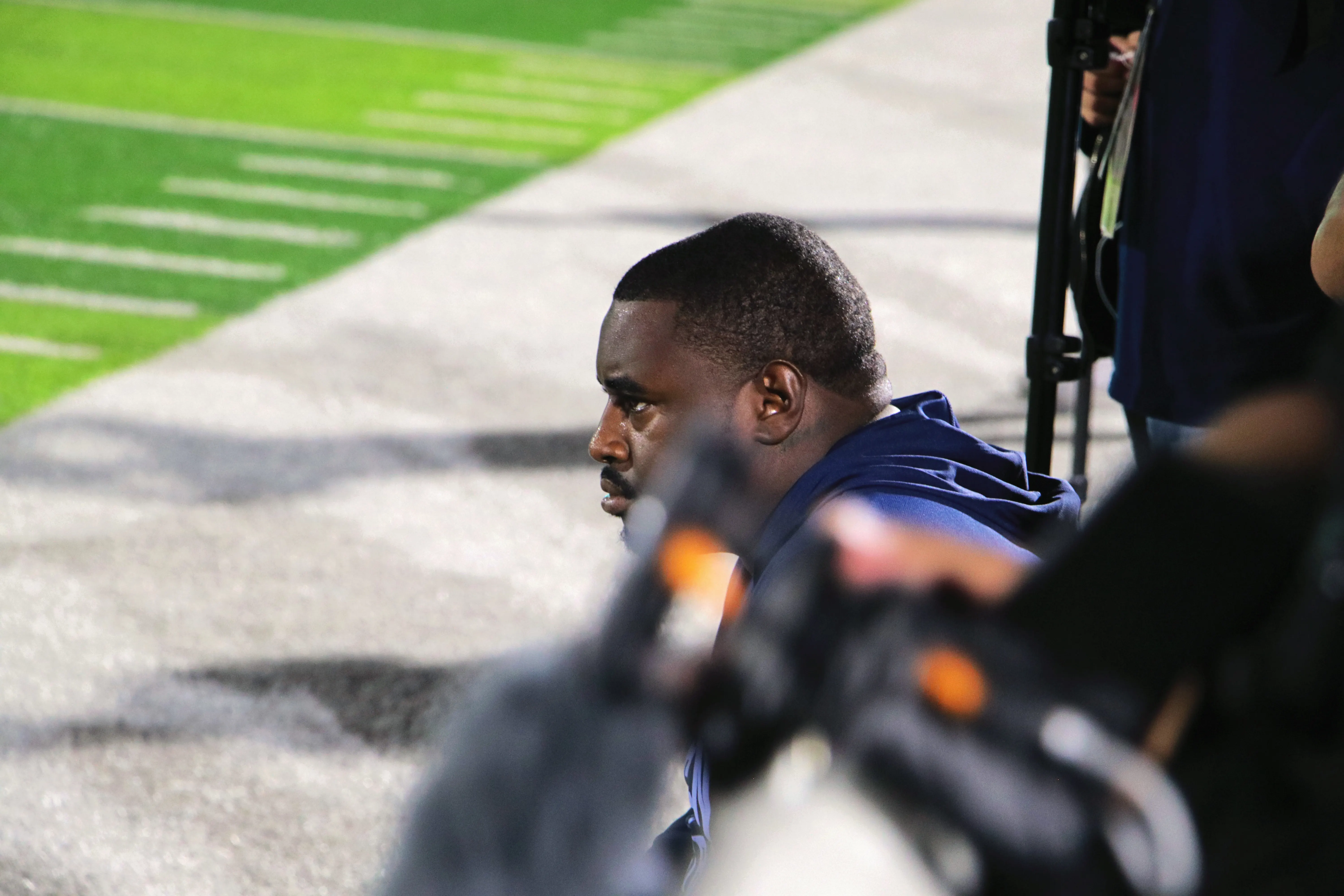 A man in a navy hoodie sits on the sideline watching the game intently, a camera rig blurred in the foreground.