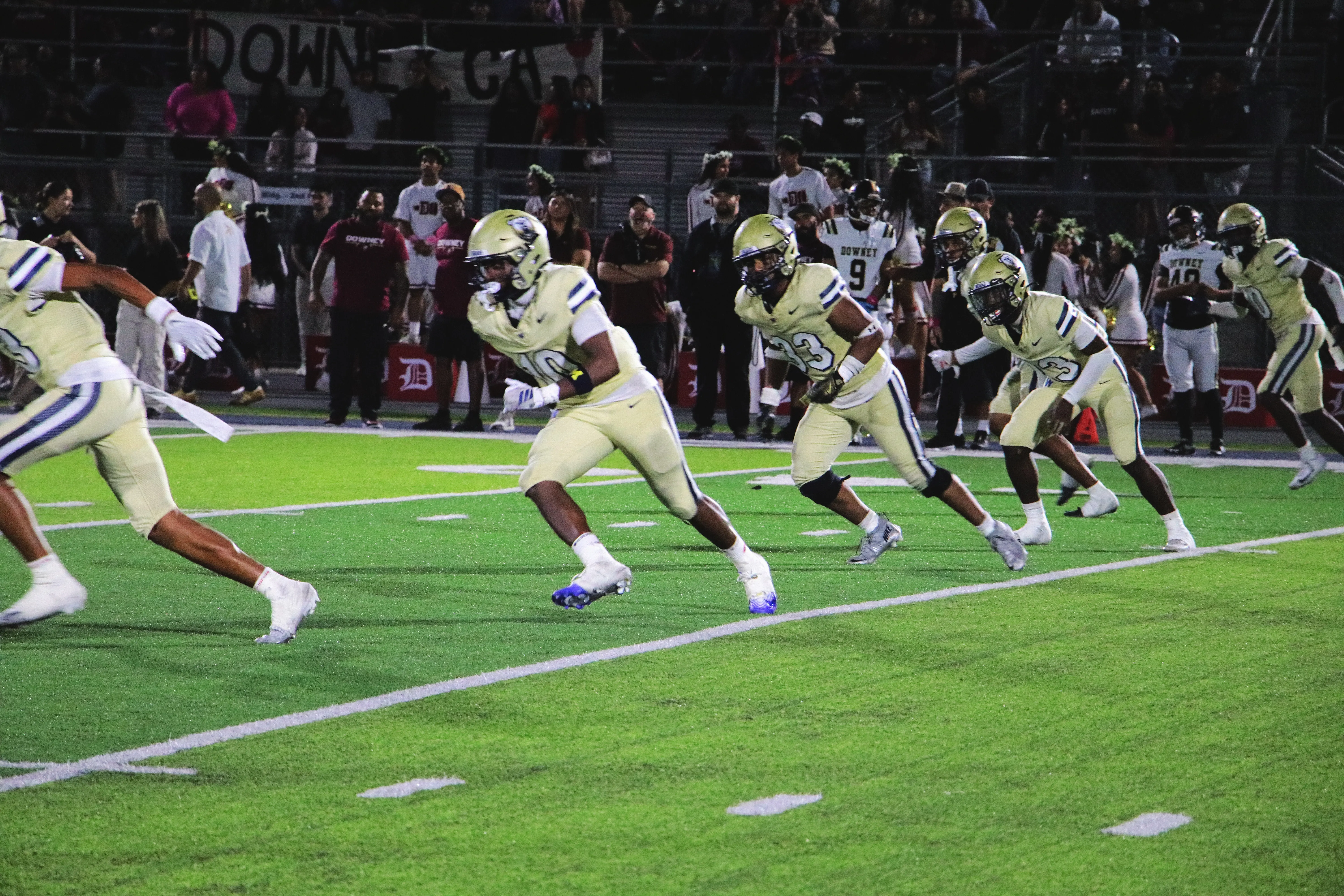 Warren Bears players in gold uniforms sprint downfield together on a green turf field, crowd lining the stands behind them.