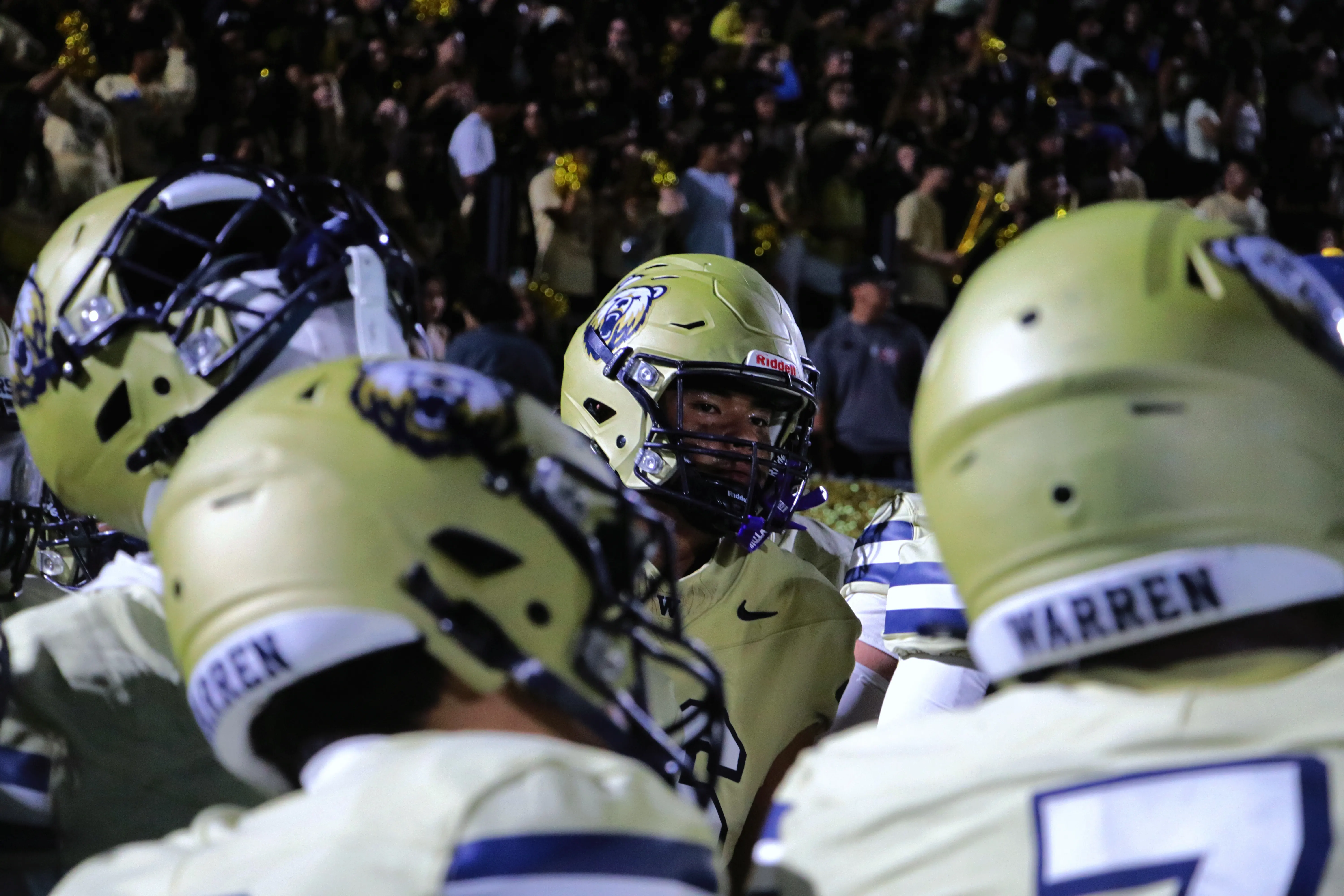 A Warren Bears player in a gold helmet looks out through a gap between teammates’ helmets, a packed crowd in the stands behind.