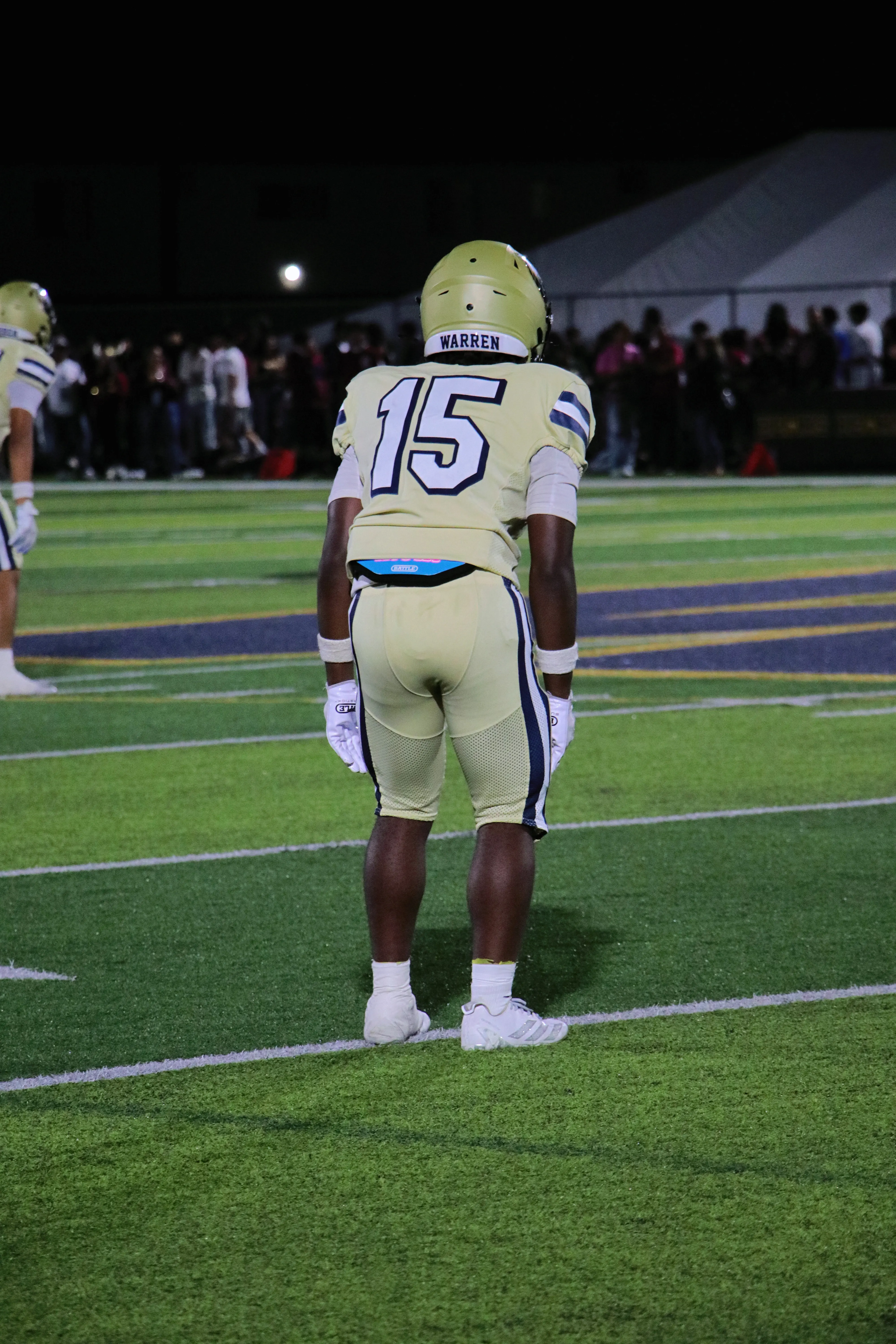 A Warren Bears player wearing number 15 stands on the field seen from behind, waiting for the next play under the night lights.